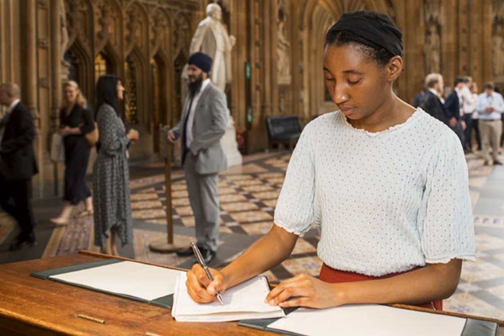 A young woman is writing with a pen on  a sheet of paper at a wooden desk on the Parliamentary estate.