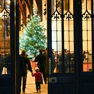 A brightly lit Christmas tree seen through the open doors of Westminster Hall. A man is holding a small boys hand and they are walking into the hall towards the Christmas  tree