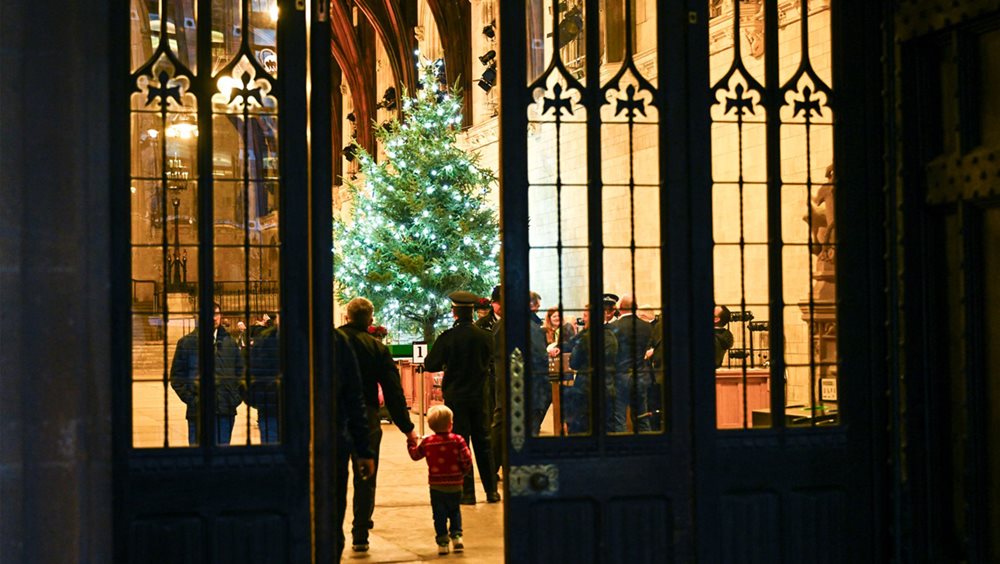 A brightly lit Christmas tree seen through the open doors of Westminster Hall. A man is holding a small boys hand and they are walking into the hall towards the Christmas  tree