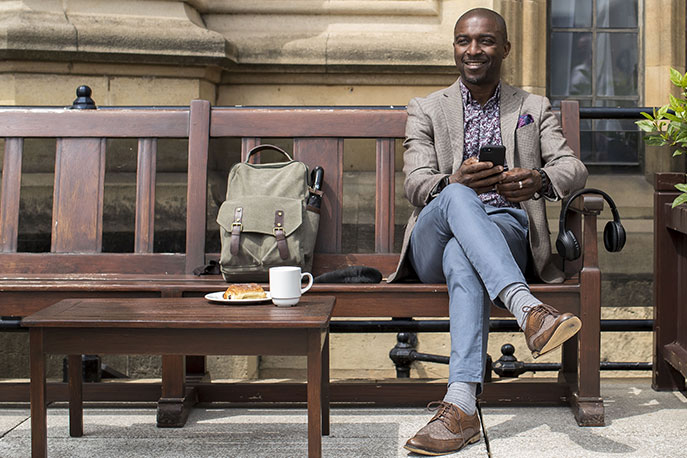 A smiling man is sitting down on a wooden bench and is holding mobile phone. 