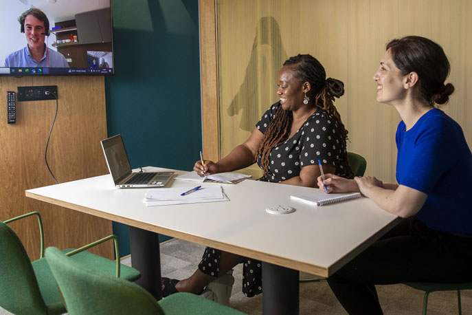 Two women sit at an office table. They are talking to a male colleague via video call.