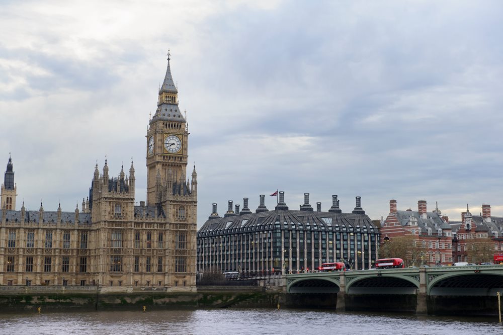 A photograph of the neo-gothic Palace of Westminster with Big Ben clock tower next to Westminster Bridge, with red buses crossing the bridge