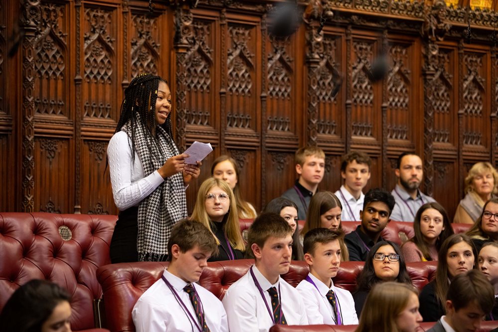 A photo of young people in the Lords chamber. A young woman is stood up and speaking to the rest of the participants.