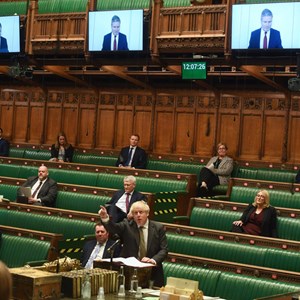 A photograph of MPs sat in the Chamber while Keir Starmer joins via videolink to question the Prime Minister during PMQs. Prime Minister Boris Johnson is pointing at the screen on which Mr Starmer appears.