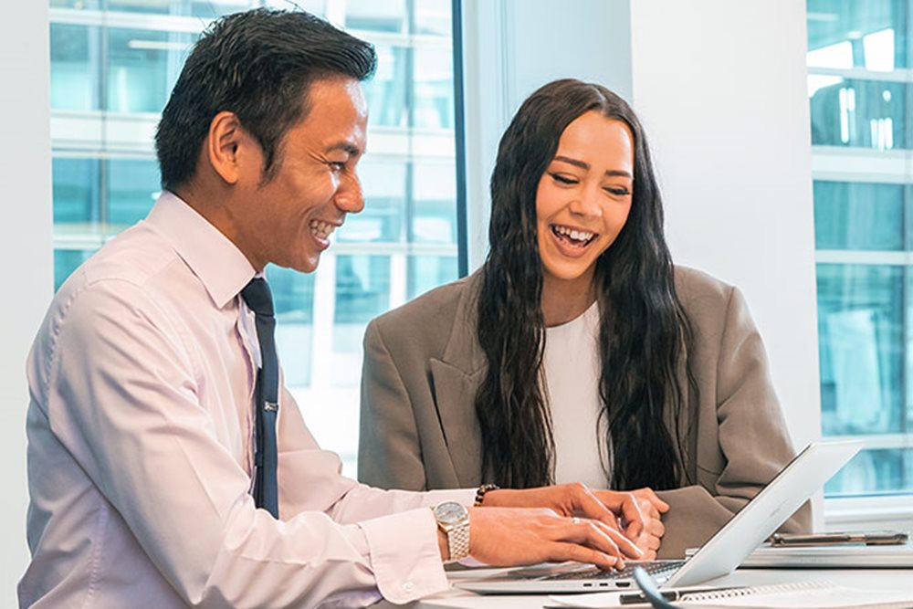 A man and a woman, both smiling, sit down at an office desk looking at a computer screen.