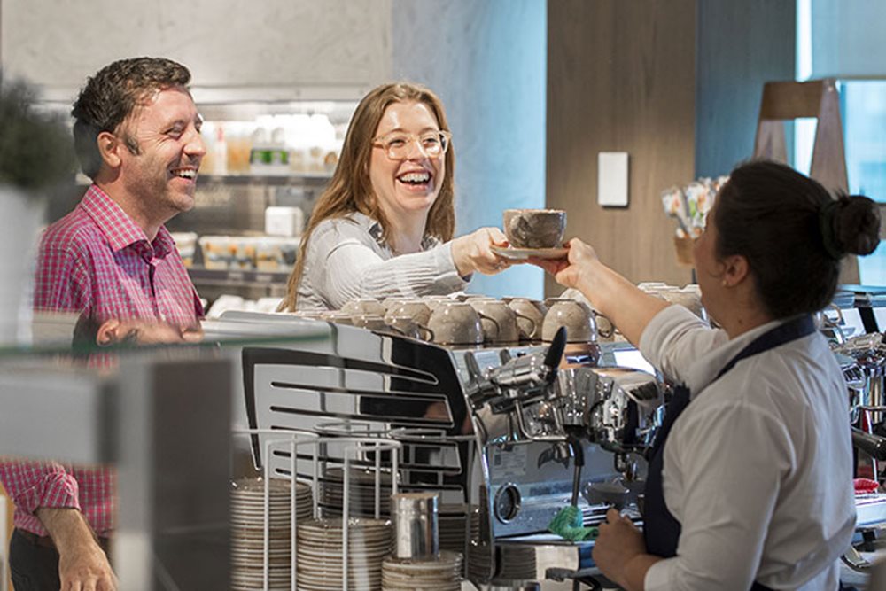 A barista is serving a coffee to a smiling female customer in the staff cafeteria. A smiling man is standing next to the female customer. 