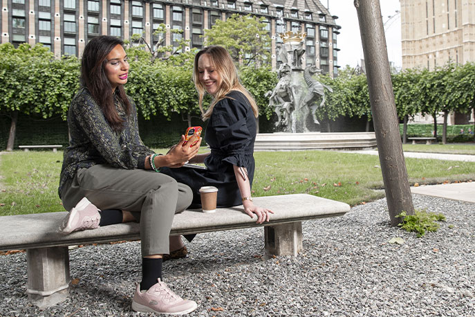 Two women are sitting on a stone bench in an urban park. They are looking at a mobile phone screen.