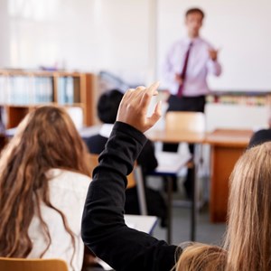 A group of students in a school classroom facing away from the camera. One student in the centre has raised their hand