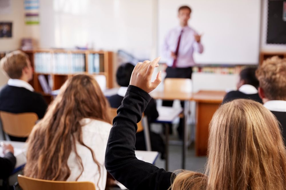 A group of students in a school classroom facing away from the camera. One student in the centre has raised their hand