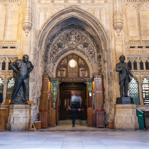 The large wooden doors of the House of Commons Chamber in the centre of the picture. blurred images of people in the centre and at either side of the door.