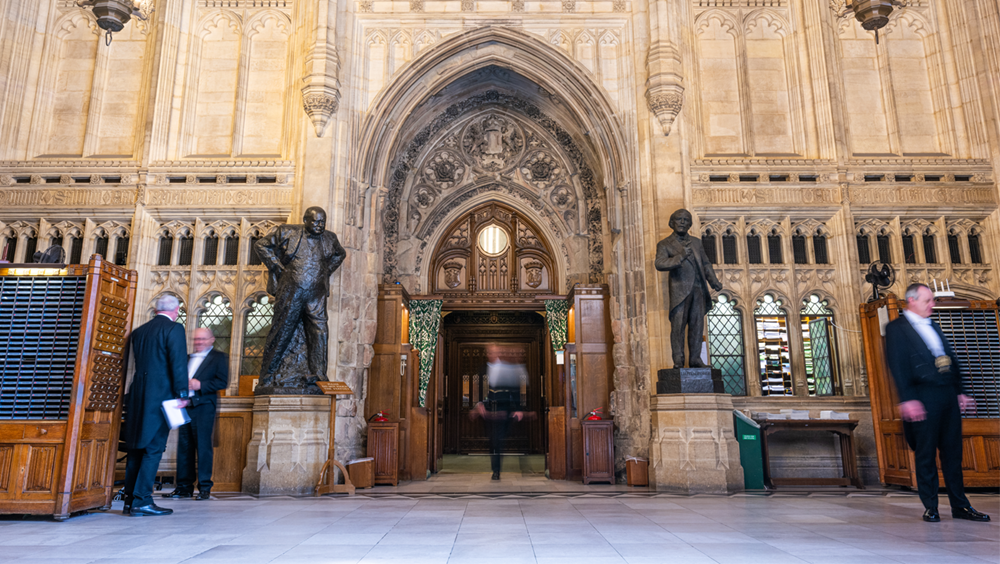 The large wooden doors of the House of Commons Chamber in the centre of the picture. blurred images of people in the centre and at either side of the door.